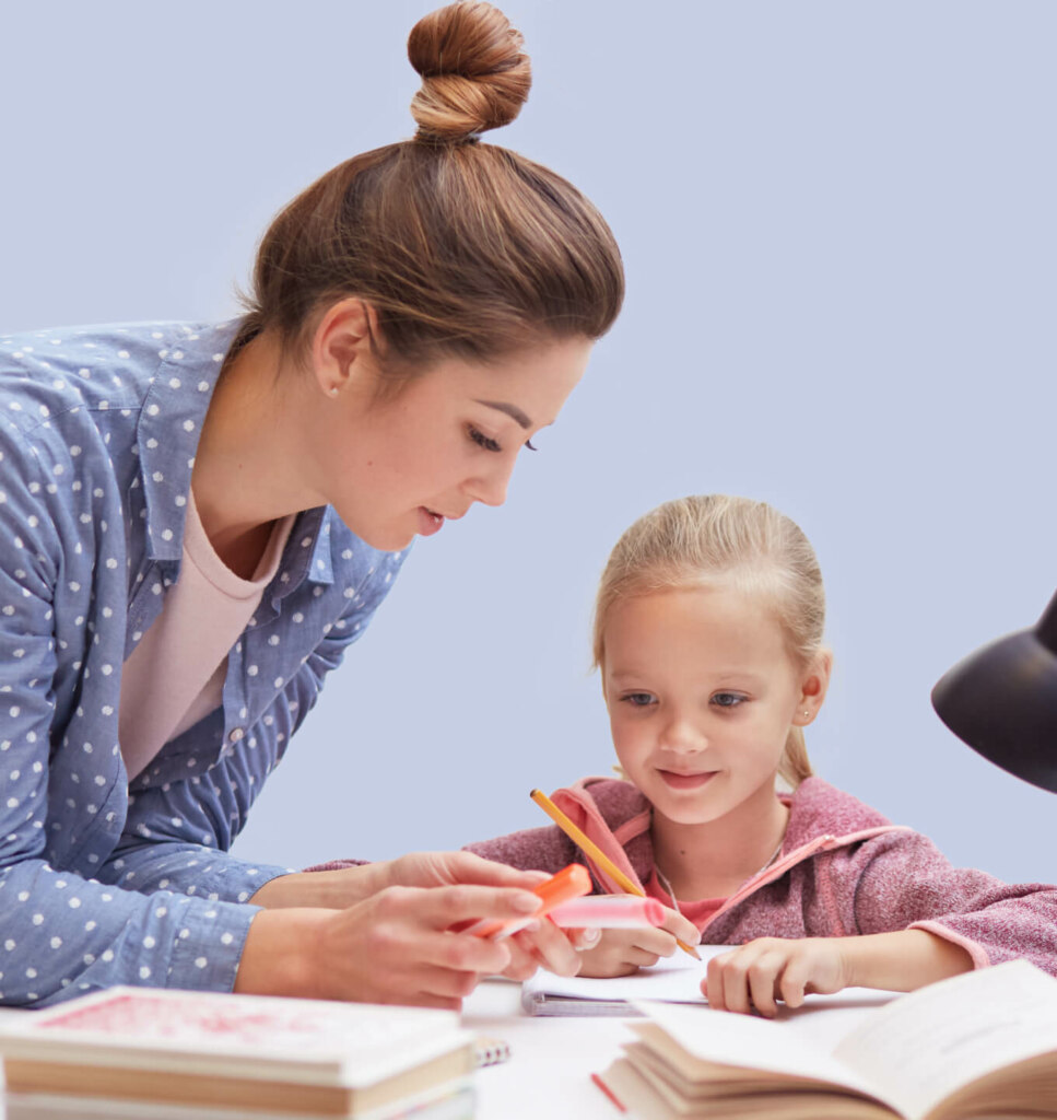 A woman is helping a young girl with her homework at a table. They are both focused on the task, with books and papers spread out around them.