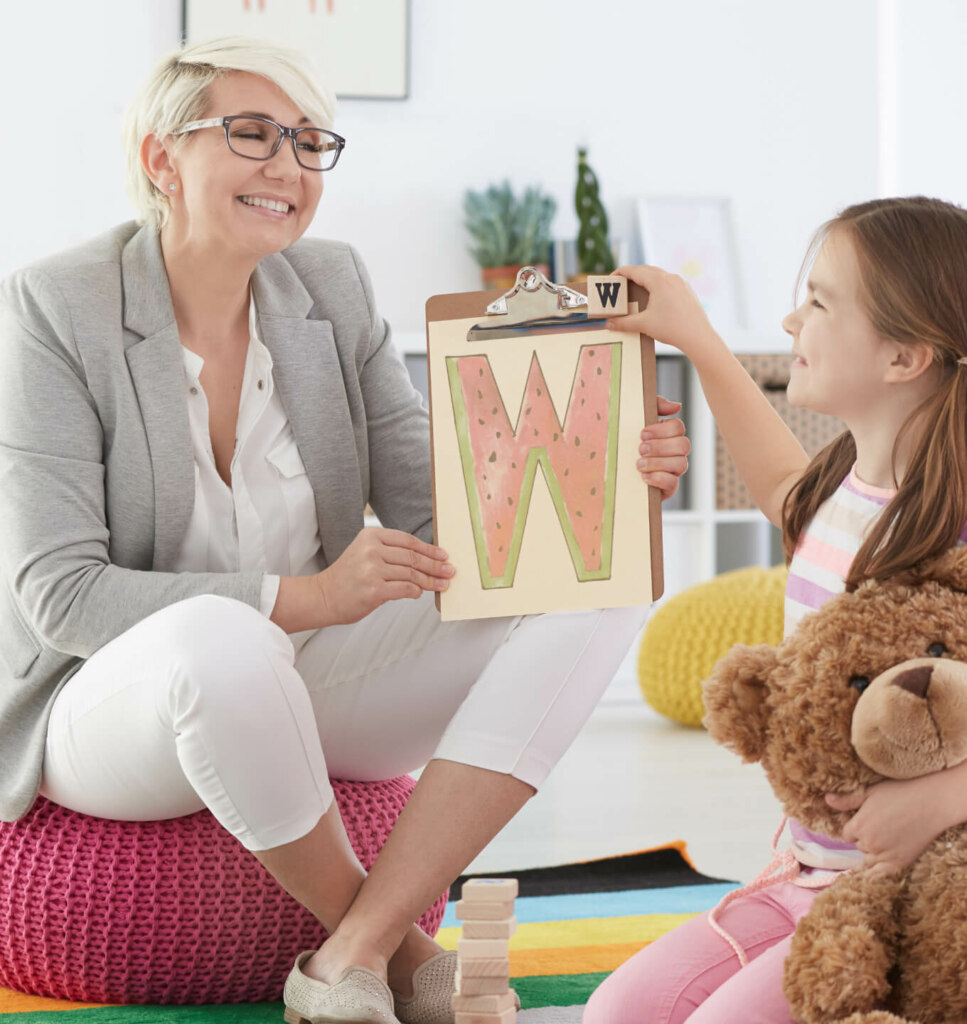 An adult sitting on a pink cushion shows a letter 'W' on a clipboard to a child holding a teddy bear. They appear to be in a brightly lit room with educational materials.