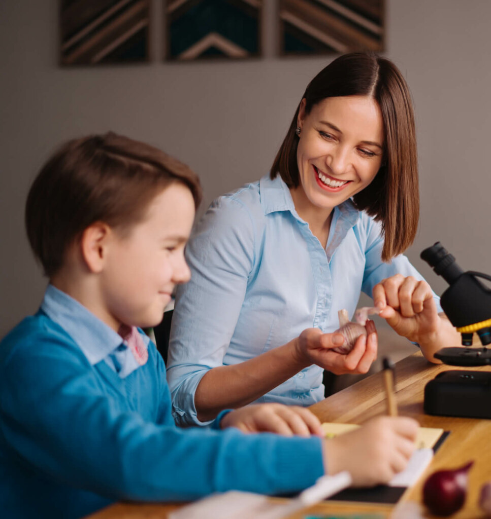 A woman in a blue shirt smiles while showing a rock sample to a young boy at a table. The boy, also in a blue shirt, is holding a pencil and writing. A microscope is on the table.