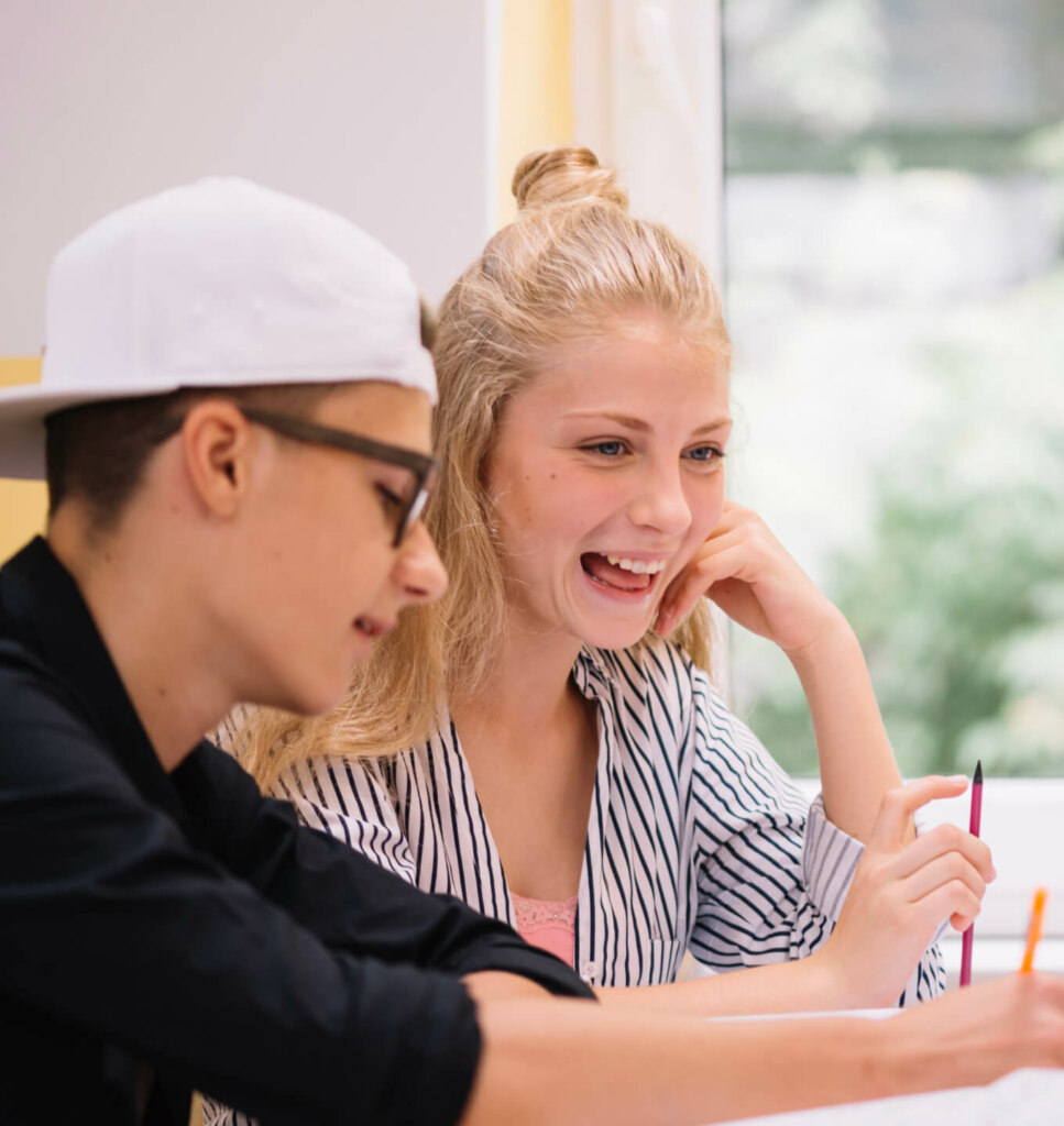 Two teenagers sit at a table, smiling and engaging in a discussion. One wears glasses and a backward cap, while the other has long hair tied up in a bun. They appear to be working on a project together.
