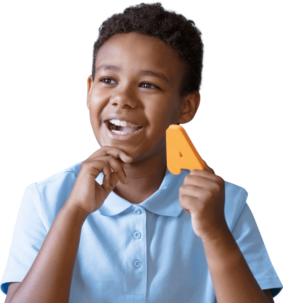 A child in a blue shirt smiling and holding a large, orange letter "A" while sitting.