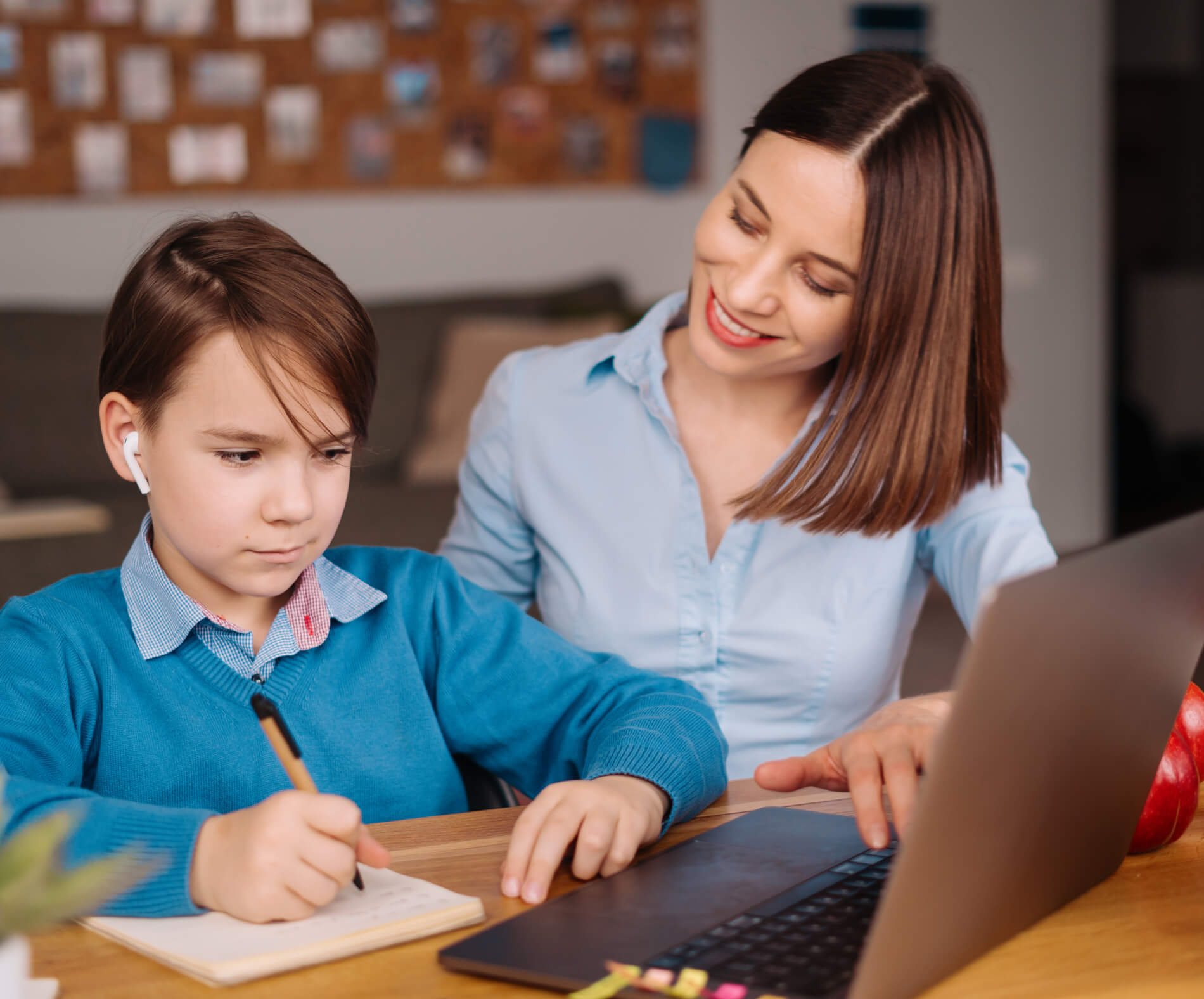A woman smiles and assists a child who is writing in a notebook while using a laptop. The child is wearing earbuds and appears focused.
